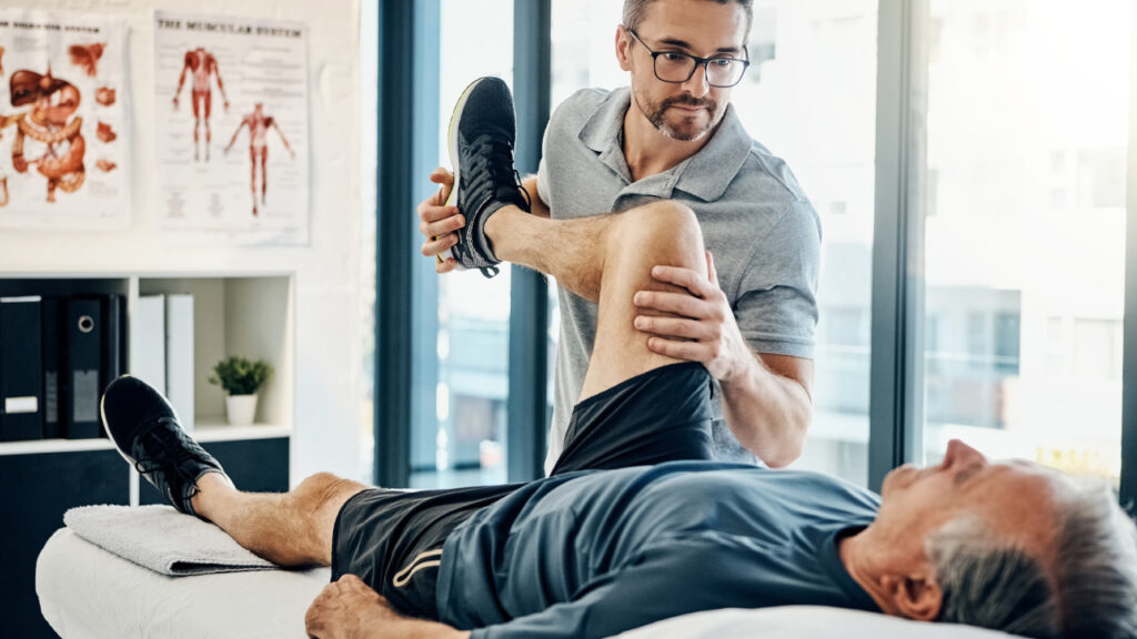 Senhor idoso deitado num consultório a fazer fisioterapia com ajuda do técnico